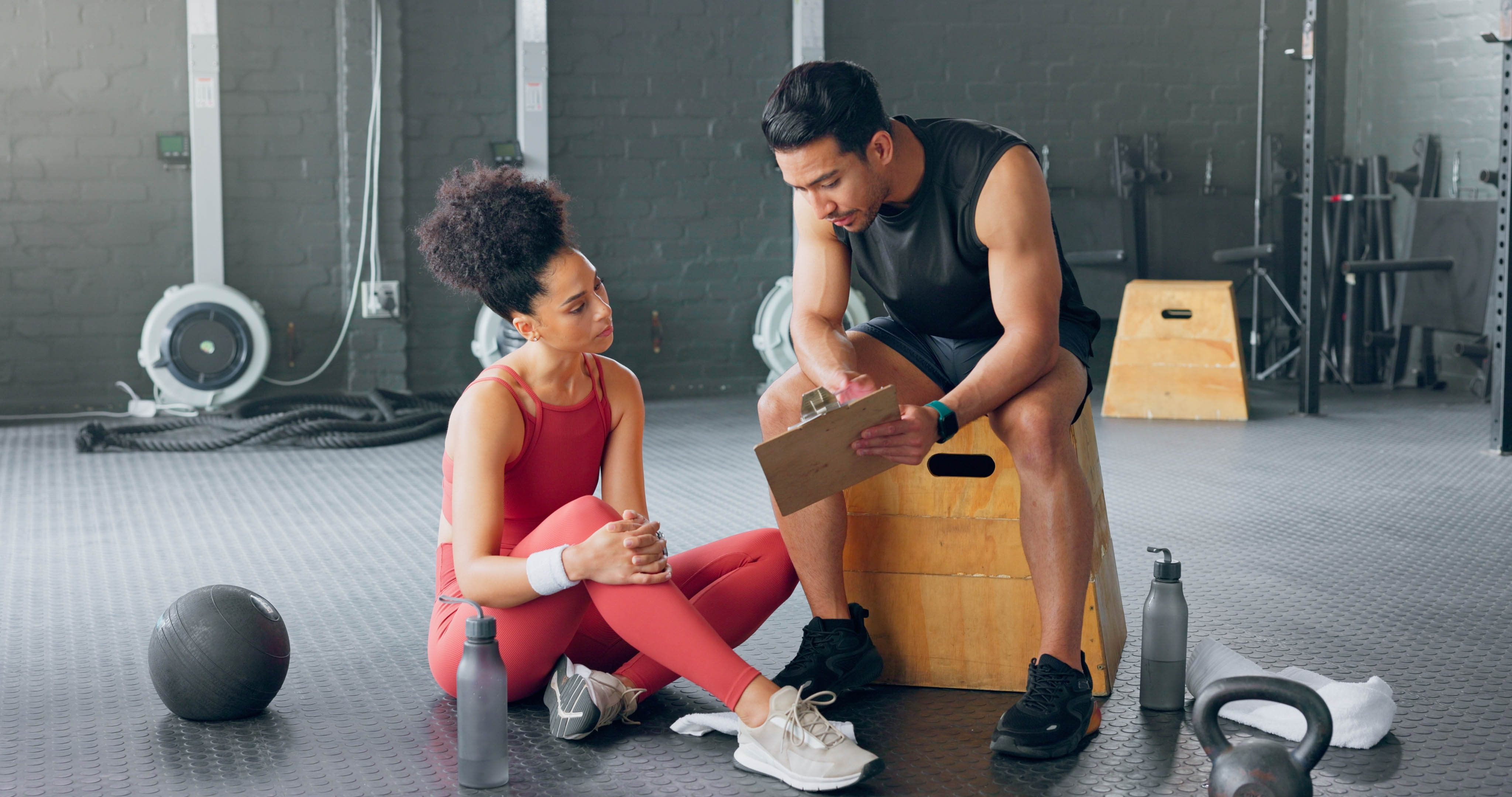 Young fit woman sitting on the ground of the gym, learning from a young male trainer who is showing her something on a clipboard